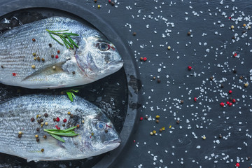 elevated view of raw fish with rosemary in tray on black table covered by salt and pepper