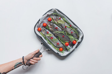 cropped image woman holding tray with fish with rosemary, bay leaves and cherry tomatoes over white table