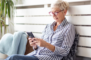 Mature woman with mobile phone on her hands sitting in room and sending messages to her friends and...