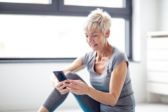 Modern short blonde hair mature woman using mobile phone while relax after exercise in corner of room with window at background