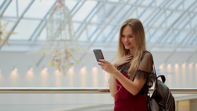Girl with a backpack talking on video coll through headphones and a mobile phone at the airport. posing for selfie on smartphone camera, hipster girl enjoying radio broadcast making photo for picture