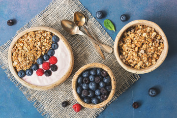 Bowl of homemade granola with yogurt and fresh berries blueberries and raspberries on blue rustic background. Healthy diet breakfast. Overhead, top view, flat lay.