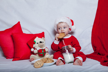 A little kid in a red cap eats a cookies and milk. Christmas photography of a baby in a red cap. New Year holidays and Christmas