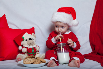 A little kid in a red cap eats a cookies and milk. Christmas photography of a baby in a red cap. New Year holidays and Christmas