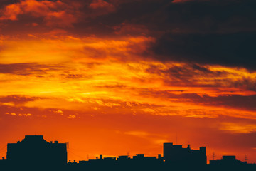 Cityscape with vivid fiery dawn. Amazing warm dramatic cloudy sky above dark silhouettes of city buildings. Orange sunlight. Atmospheric background of sunrise in overcast weather. Copy space.