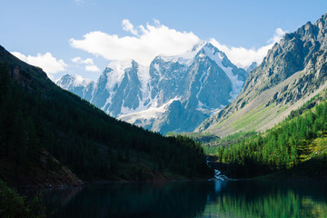 Fototapeta na wymiar Amazing glacier under blue sky. Forest reflected on clean water of mountain lake. Huge cloud on giant wonderful snowy mountains. Rich vegetation of highlands. Atmospheric landscape of majestic nature.