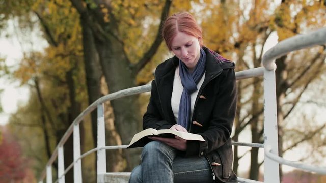 A young woman reads the Bible in a public park by autumn.