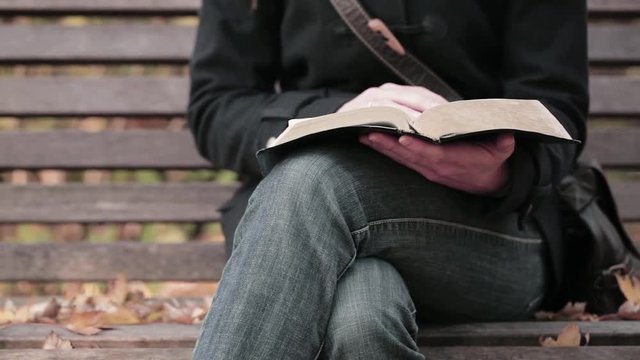 A young woman reading the Bible on a bench in a park.