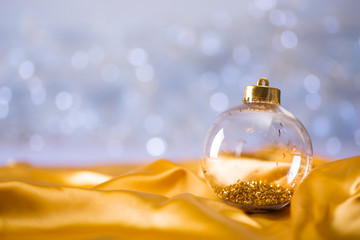 Glass transparent Christmas ball with gold tinsel inside on golden satin fabric on light bokeh...