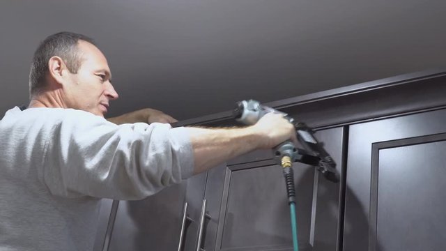 Construction worker using brad nail air gun to Crown Moulding on white kitchen wall cabinets framing trim, with the all power tools