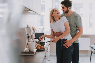 Cooked with love. Cute laughing blonde frying something in skillet. Her bearded husband is hugging...