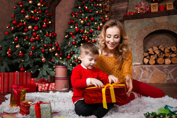 Fototapeta na wymiar Child kid boy in red warm sweater with young mother female packed gift present for his parent mother and father during winter holiday at decorated home. Merry Christmas and Happy New Year concept.