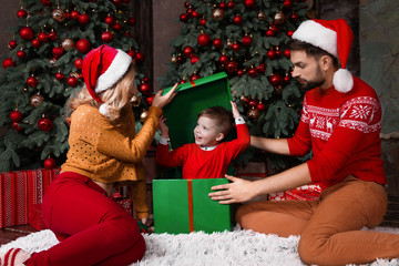 Happy young family mother and father wear santa hats got gift's boy son in green gift's box near decorated christmas tree at home during winter holidays. Merry Christmas and Happy New Year concept,