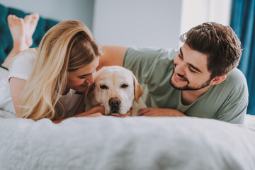 Optimistic young couple lying in bed while having fun with their dog