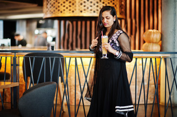 Pretty indian girl in black saree dress posed at restaurant with orange juice at hand.