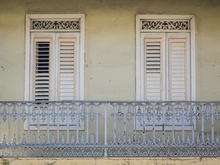 A fancy balcony railing in front of two shuttered windows with carved trim in an old Caribbean building