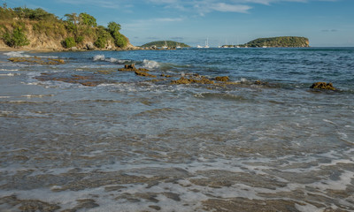 Fototapeta premium View from the water of waves coming into shore with rocky headlands, sailboats, and islands along the horizon on a sunny day in the Caribbean.