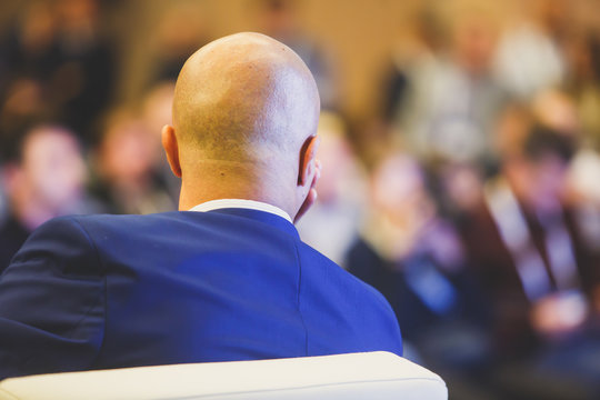 A back view of a male Speaker in a blue suit giving a talk on a Stage at a corporate Business Conference in front of Audience at the Conference hall, economic event