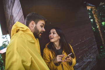 Look here. Waist up portrait of young man and woman in raincoats lively discussing some information from mobile phone. Small wooden house on background