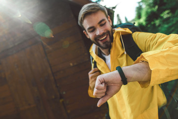 Being in time in journey and adventures. Close up portrait of bearded cheerful male traveler looking at his watch near wooden house. Copy space on left