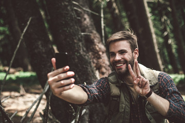 Have a rest and connect the world. Waist up portrait of happy male traveler showing peace gesture while making selfie in green forest