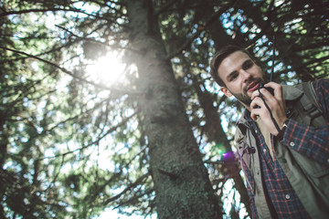 Fototapeta na wymiar Concept of experienced journey and connection. Low angle portrait of cheerful bearded man talking on portable radio set while walking in green sunny forest