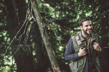 Concept of interesting journey and adventure. Waist up portrait of cheerful bearded male walking uphill in green forest. Copy space on left