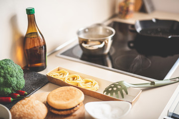 Close up of desk with tagliatelle, hamburgers, vegetables and large spoon for draining spaghetti. Pot and frying pan on modern stove