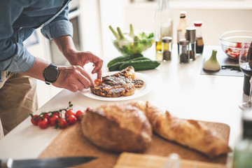 Close up of male hands holding slice of tomato near plate with grilled meat. Gentleman standing near white kitchen table