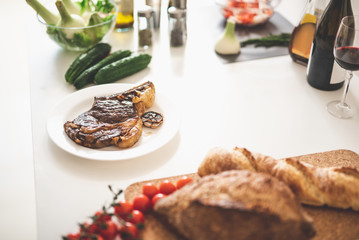 Bon appetite. White kitchen table with grilled meat, glass of wine, cucumber, onion and tomatoes