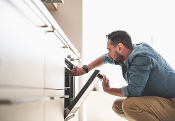 Side view portrait of handsome gentleman in denim shirt looking into oven while holding tongs. He is squatting on the floor