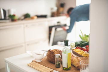 White kitchen desk with bread, olive oil, spice, pasta and sauce boat. Gentleman checking oven