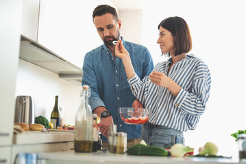 Waist up portrait of charming woman distracting bearded from cooking. Couple standing near kitchen...