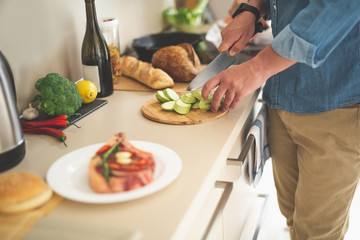 Close up of male arms chopping ingredients with knife. Gentleman standing near kitchen table with vegetables, bottle of wine, beef steak and bread