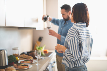 Back view portrait of charming woman standing in kitchen and staring at bearded man while he preparing drink for romantic dinner. Focus on girl