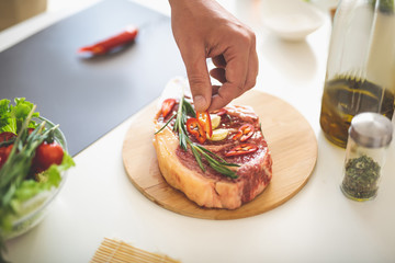 Close up of male hand holding slice of chili pepper while putting it on raw beef steak with garlic and rosemary