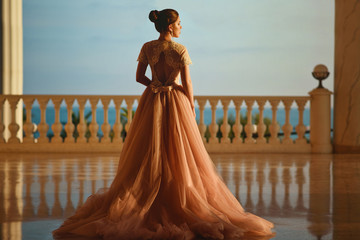 Beautiful woman in luxurious ballroom dress with tulle skirt and lacy top standing on the large balcony with sea view. Back view - Powered by Adobe