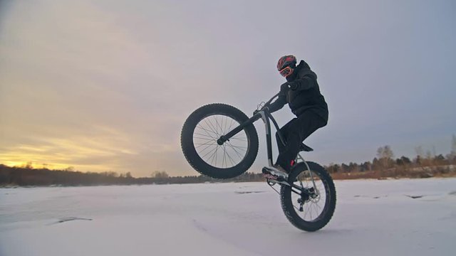 Professional extreme sportsman biker riding fat bike in outdoors. Cyclist ride in winter snow forest. Man does trial trick pedal kick jump on mountain bicycle with big tire in helmet and glasses.