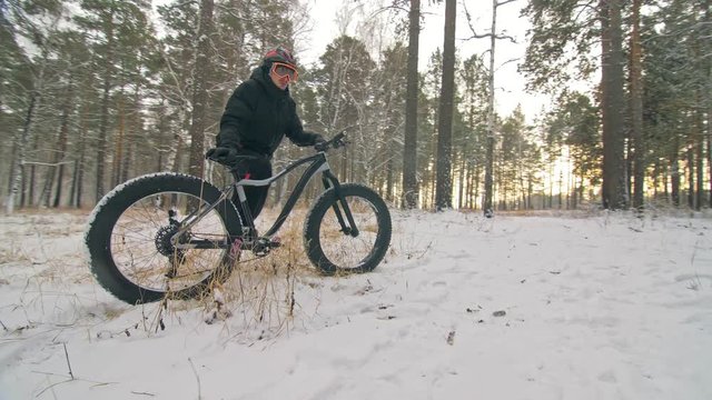 Professional extreme sportsman biker stand a fat bike in outdoors. Cyclist recline in the winter snow forest. Man walk with mountain bicycle with big tire in helmet and glasses. Slow motion in 60fps.
