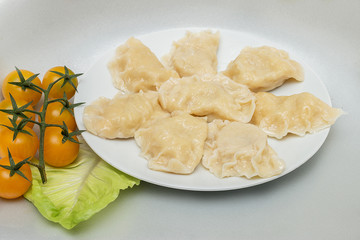 Dumplings lie on a white plate. On a gray background is a plate with dumplings and beside it are yellow tomatoes and a leaf of lettuce.