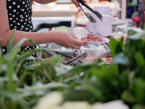 Woman's hands taking pork from tray at a buffet hotpot ( Moo Joom ) restaurant with a defocus vegetables in the foreground