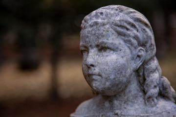 Fototapeta na wymiar Partial View of Victorian Statuary with a Girl's Face and Shoulders in Soft Focus with Out of Focus Foliage and Trees in Background