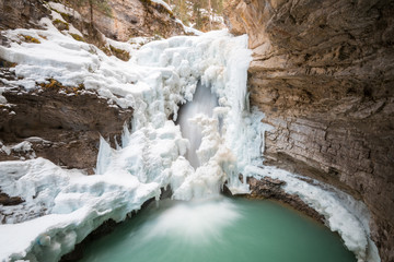 A half frozen waterfall flows into a green pool surrounded by huge ice masses from a cold winter