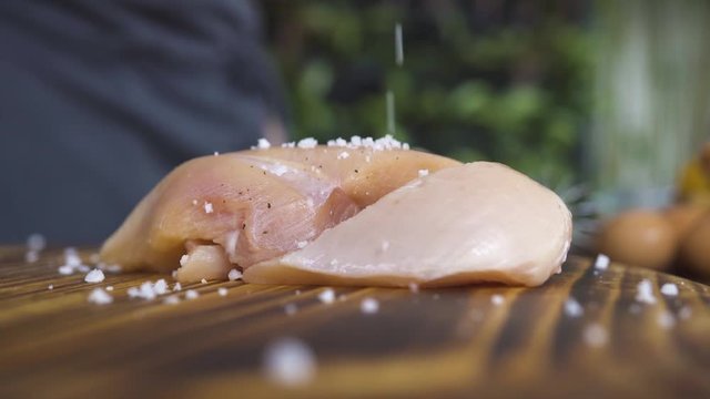 Salt pouring on chicken breasts on kitchen board close up. Raw fresh chicken fillet marinating on wooden table. Process preparation marinated meat for bbq. Cooking food and meat dish.