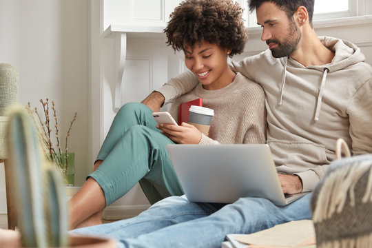 Image of interracial lovely couple in love rest at home interior, satisfied with communication and gadgets, drink coffee, hug warmly, view common photos on cell phone, pose in modern flat, chat online