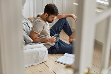 Fototapeta premium People, literacy, spare time concept. Focused unshaven man enjoys fresh beverage during day, focused in book, reads scientific literature, prepares for final examination sits on floor. View from doors