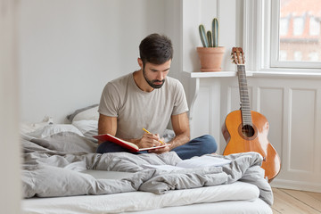 Photo of serious man with dark bristle, writes down story in diary about what happened during day,...