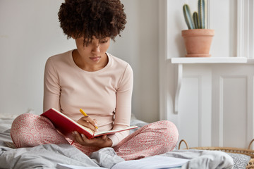 Photo of attractive black lady in casual clothes writes story into notebook with pen, keeps legs...