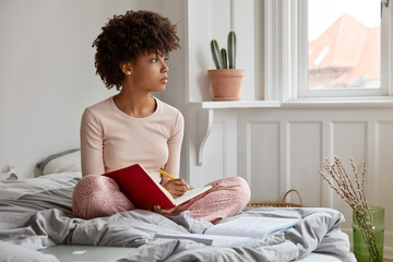 Thoughtful dark skinned young woman writes at textbook, sits in lotus pose, wears nightwear, sits...