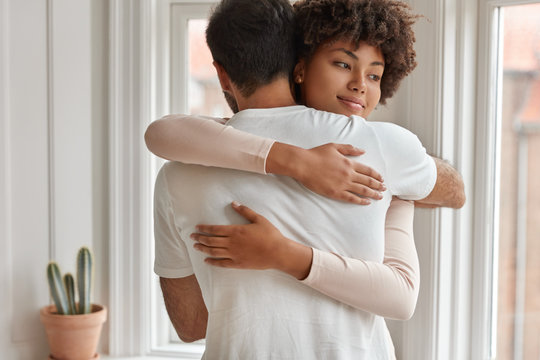 Happy mixed race couple embrace each other, express support and love, have friendly relationships, pose near window in living room, enjoy togetherness. Diverse boyfriend and girlfriend hug indoor